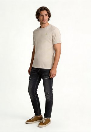 Mann steht, trägt beige kurzärmeliges T-Shirt, schwarze zerrissene Skinny-Jeans und braune Turnschuhe auf weißem Hintergrund.