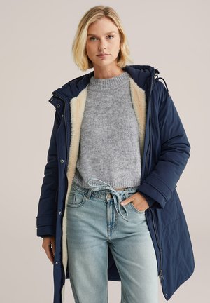 Parka marine avec une fermeture éclair à l'avant et des boutons-pression ; doublé de fausse fourrure claire ; porté par-dessus un pull en tricot gris et un jean bleu clair.
