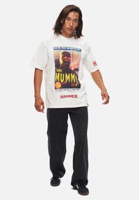 T-shirt blanc à manches courtes avec un graphique vintage du film "La Momie". Un pantalon noir ample et des baskets noires complètent la tenue.