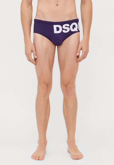 Uomo in piedi a piedi nudi che indossa slip da nuoto viola con grande testo bianco "DSQU" sulla parte anteriore, in un contesto di sfondo semplice.