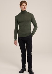 Zeleni pulover s turtleneck ovratnikom, prilegajoče zasnove in dolgimi rokavi, v kombinaciji s črnimi ozkimi hlačami in črnimi gležnjarji. Gladka tekstura.