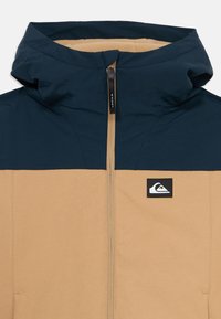 Mikina s kapucňou, ktorá má tmavomodrú hornú časť, pieskovú dolnú časť, predný zips a logo Quiksilver na ľavej hrudi.
