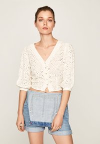 Blusa corta bianca con motivo ad occhio di pernice, scollatura a V e chiusura con bottoni, abbinata a pantaloni corti in denim chiaro con una tasca decorativa.