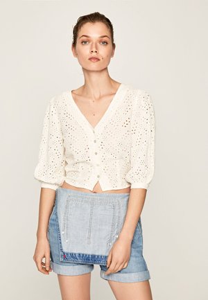 Blusa corta bianca con motivo ad occhio di pernice, scollatura a V e chiusura con bottoni, abbinata a pantaloni corti in denim chiaro con una tasca decorativa.