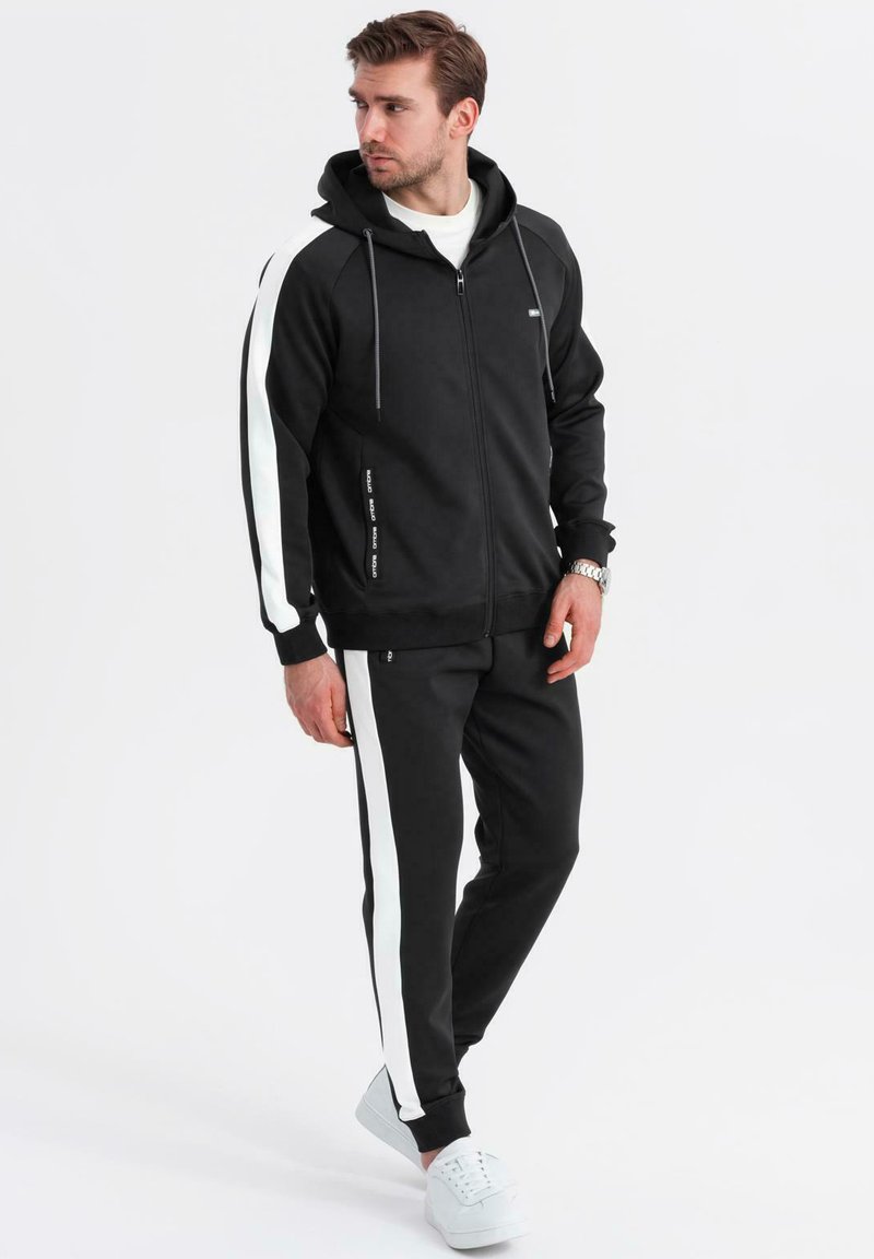 Uomo con felpa nera con zip e pantaloni coordinati con strisce laterali bianche, indossa scarpe da ginnastica bianche, in piedi e guarda verso sinistra.