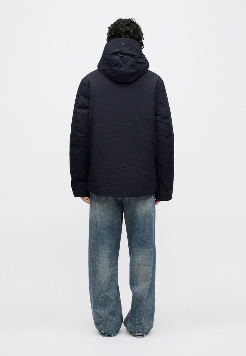 Navy blue, waterproof jacket with a hood; features a smooth texture, elastic cuffs, and a slightly longer back design. Worn with light blue jeans.