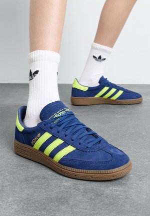 Baskets en daim bleu avec trois rayures fluorescent vert, semelle en caoutchouc brun texturé et accents assortis. Chaussettes blanches avec logo noir.