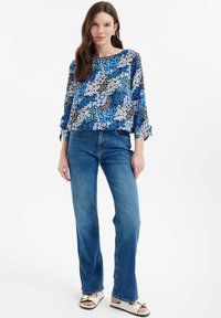 Blusa floreale con colori blu, nero e bianco, scollatura tonda e maniche 3/4 con dettagli a fiocco. Indossata con jeans in denim a gamba dritta.