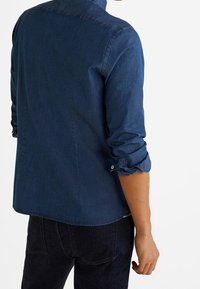 Uomo che indossa una camicia di jeans blu scuro con le maniche arrotolate e jeans neri, in piedi con la schiena parzialmente voltata.