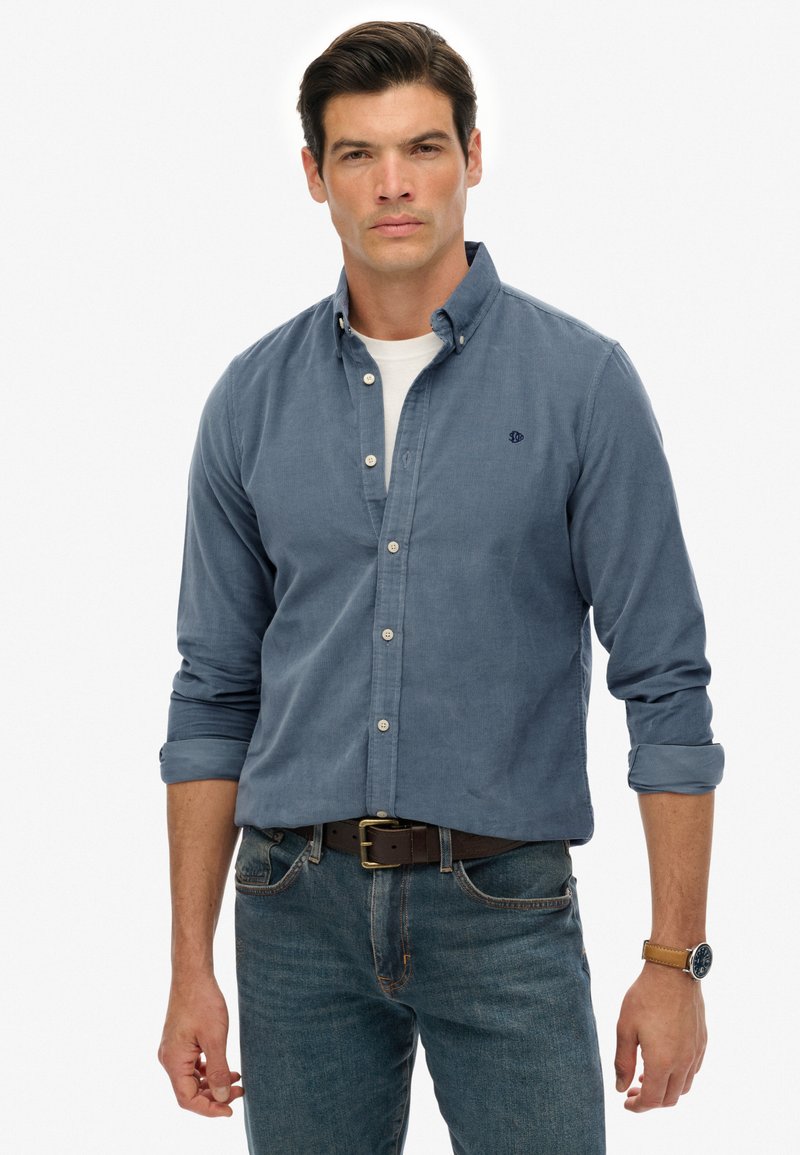 Uomo con camicia blu a bottoni sopra una maglietta bianca, jeans blu con cintura marrone e un orologio al polso sinistro, in piedi contro uno sfondo bianco.