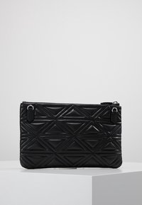 Černá prošívaná kabelka s geometrickými trojúhelníkovými vzory a horním zipem, vystavená na bílém podstavci na neutrálním pozadí.