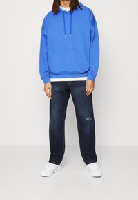 Sudadera con capucha azul y bolsillo delantero, combinada con jeans azul oscuro con detalles desgastados y zapatillas blancas.