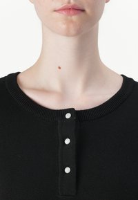 Black ribbed sweater with a round neckline and three silver button accents down the front. Close-up view focuses on the neckline area.