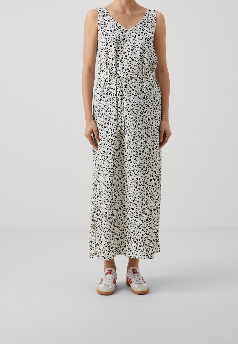 Maxi-jurk in crème met blauw dierenprint, mouwloos ontwerp, V-hals, cinched taille, gecombineerd met witte sneakers met rode accenten.