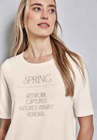 T-shirt crème à manches courtes avec un texte graphique en marron : « L'ART PRINTANIER CAPTE LE RENOUVEAU VIBRANT DE LA NATURE. » Texture lisse.