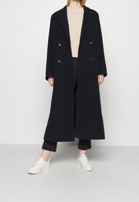 Granatowy wełniany płaszcz z podwójnym zapięciem, klapami i bocznymi kieszeniami, noszony na beżowej bluzce i ciemnych spodniach, w połączeniu z białymi sneakersami.