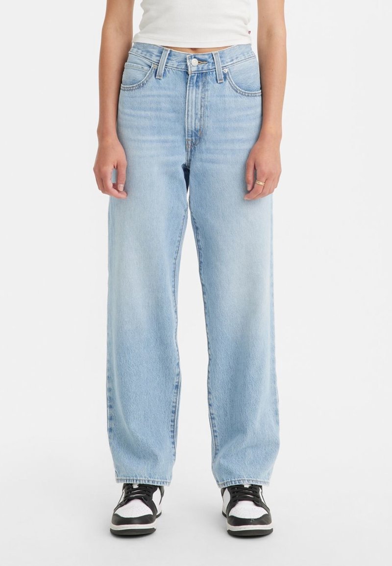Personne portant un jean bleu clair taille haute à jambes larges, un top court blanc et des baskets noires et blanches, debout sur un fond uni.