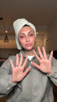 Giovane donna con un asciugamano avvolto sulla testa mostra entrambe le mani con unghie a punta francese, indossando una giacca grigia con zip all'interno.