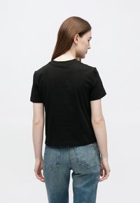 Femeie cu păr lung și castaniu, purtând un tricou negru cu mâneci scurte și blugi albaștri, cu fața spre spate pe un fundal simplu și deschis la culoare.