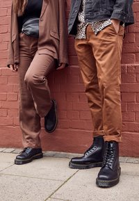 Bottes en cuir noir avec des coutures jaunes, associées à un pantalon marron et un blazer assorti sur une personne, et une veste en denim noire avec une chemise à motifs sur une autre.