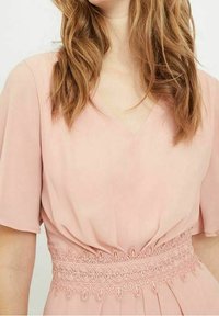 Robe rose clair avec des manches courtes, décolleté en V et des détails en dentelle complexes à la taille. Texture de tissu lisse et fluide.