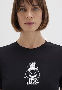 Black cotton t-shirt featuring a white graphic of a cat on a jack-o'-lantern with the text "STAY SPOOKY" below. Short sleeves and round neck.