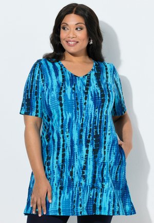 Kobieta o długich ciemnych włosach nosi niebiesko-czarną tunikę z krótkim rękawem w wzory, uśmiecha się i patrzy na bok, mając jedną rękę w kieszeni.