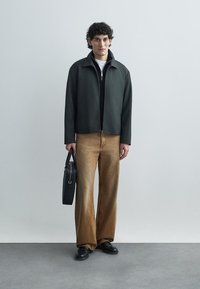 Tmavozelená bunda na zips cez čierne tričko, v kombinácii s voľnými hnedými nohavicami. Drží čiernu tašku, má na sebe čierne topánky. Hladká textúra.