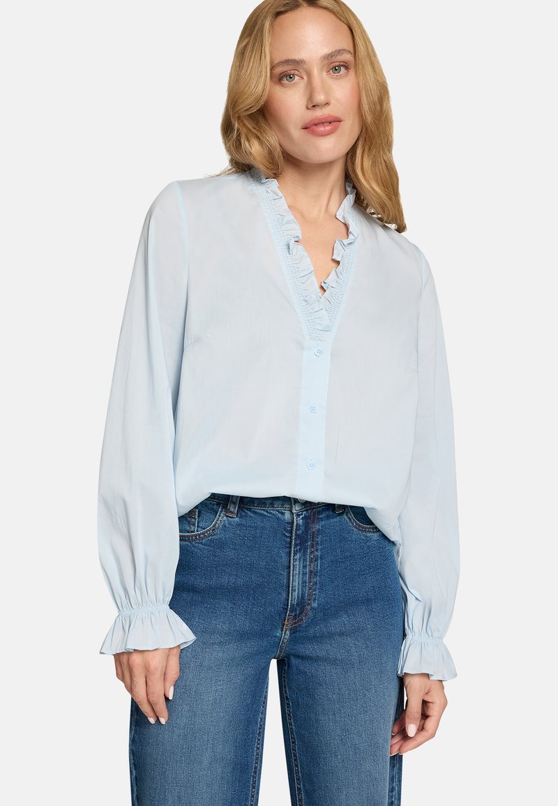 Blouse bleu clair avec encolure à volants, manches longues avec poignets à volants, et fermeture boutonnée devant, associée à un jean en denim foncé.