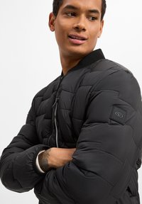 Black puffer jacket with a quilted texture, ribbed collar, and zippered front. Features a logo patch on the sleeve.