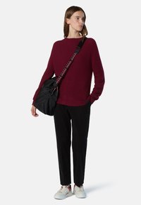 Maglione tricot bordeaux con maniche lunghe, borsa nera con tracolla decorata, pantaloni neri e sneakers bianche con dettagli rossi.