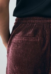 Bordo aksominiai kelnių su elastine juosta ir užpakaline kišenė. Audinys turi švelnų, tekstūruotą išvaizdą ir lygią paviršiaus apdailą.