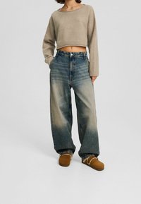 Beżowy krótki sweter, dżinsy z wysokim stanem i szerokimi nogawkami z efektownym przejściem koloru od jasnego do ciemnego, w zestawieniu z brązowymi butami wsuwanymi.