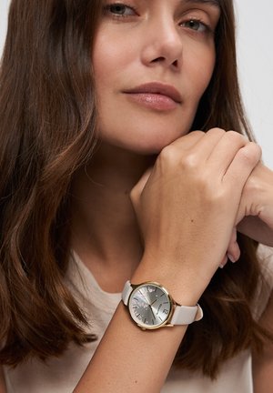 Frau mit langen braunen Haaren, die das Kinn auf die Hand stützt, trägt eine weiße Uhr mit goldenem Rand und floralem Design auf dem Zifferblatt.