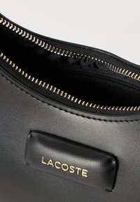 Fekete bőr Lacoste neszesszer arany cipzárral, részben nyitva, fekete szövetbelsővel és az elején varrott márkajelzéssel.