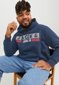 Sudadera navy con logo frontal "JACK & JONES", cordones y bolsillo tipo canguro, combinada con jeans de mezclilla azul claro. Textura suave, corte casual.