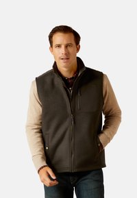 Schwarze Fleece-Weste mit hohem Kragen und Reißverschlusstasche, über beigefarbenen Langarmshirts getragen. Mit einem Frontverschluss und Seitentaschen ausgestattet.