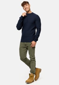 Marineblå tekstureret sweater, olivengrøn slim-fit bukser og lysebrune snøresko med slidstærke såler. Modellen poser med et afslappet udtryk.