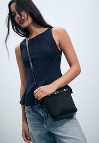 Giovane donna che indossa un top senza maniche a righe blu scuro e jeans azzurri, mentre tiene una piccola borsa a tracolla nera contro uno sfondo chiaro.