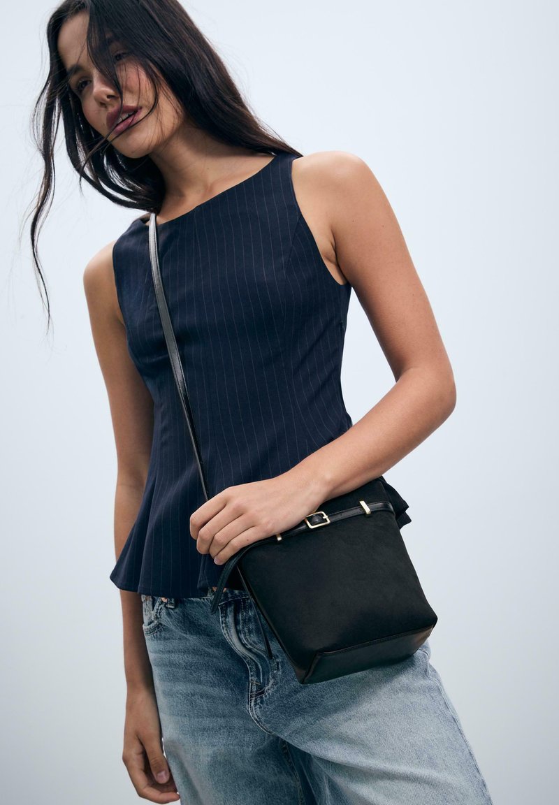 Giovane donna che indossa un top senza maniche a righe blu scuro e jeans azzurri, mentre tiene una piccola borsa a tracolla nera contro uno sfondo chiaro.