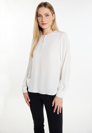 Blonde woman wearing a long-sleeve white blouse with a small front keyhole detail and black pants, standing against a white background.