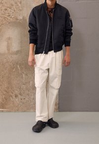 Black bomber jacket with zipper, white cargo pants, and black clogs. Smooth fabric with a soft texture, featuring pockets and a logo detail.