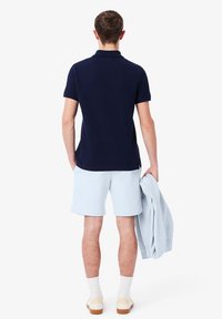Polo shirt bleu marine, shorts bleu clair, tenant un sweat-shirt gris. Chaussettes blanches et chaussures crème. Vue de dos, design minimaliste, sans motifs.