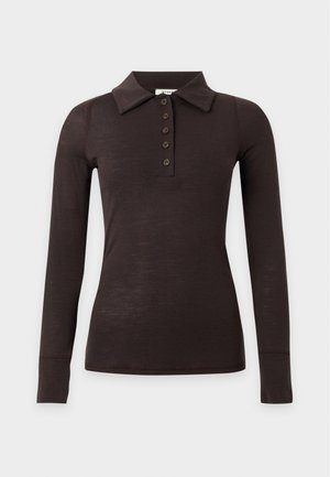 Mujer con camisa negra de manga larga con cuello y pantalones crema de pierna ancha, de pie con las manos en los bolsillos contra un fondo blanco liso.