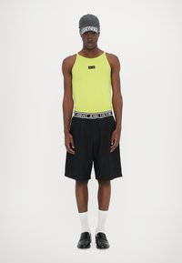 Neon grønn tanktop og svarte shorts med en merket midje, kombinert med hvite sokker og svarte loafers, toppet med en grå caps.