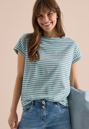 Uśmiechnięta kobieta w zielono-białej paskowanej koszulce z krótkim rękawem i niebieskich dżinsach, trzyma jasnoniebieską poduszkę na beżowym tle.