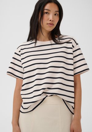 Jeune femme aux longs cheveux foncés portant une chemise blanche à manches courtes avec des rayures horizontales noires et un pantalon clair.