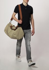 Beżowa torba podróżna z ciemnobrązowymi paskami, gładką fakturą, zaokrąglonym kształtem i bocznymi uchwytami; noszona przez osobę w codziennym ubraniu.