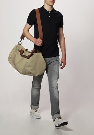 Béžová taška typu duffel s tmavě hnědými popruhy, hladkou texturou, zaobleným tvarem a bočními rukojeťmi; nošená osobou v neformálním oblečení.