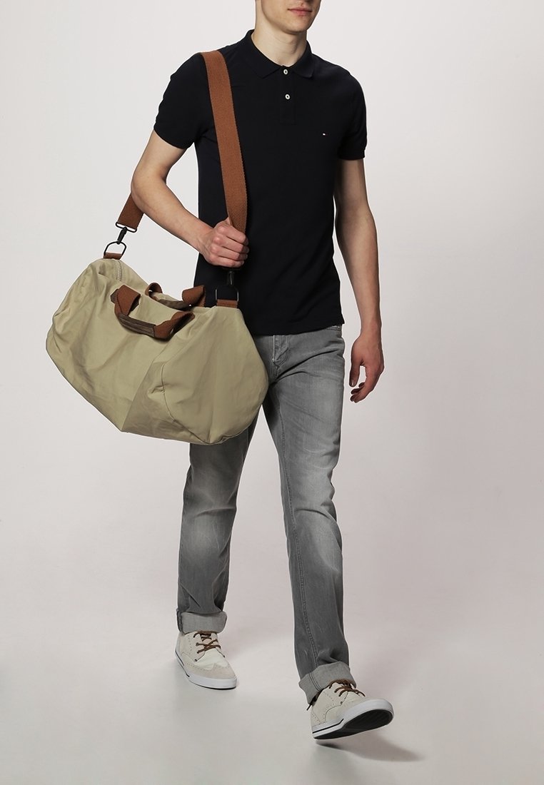 Beżowa torba podróżna z ciemnobrązowymi paskami, gładką fakturą, zaokrąglonym kształtem i bocznymi uchwytami; noszona przez osobę w codziennym ubraniu.